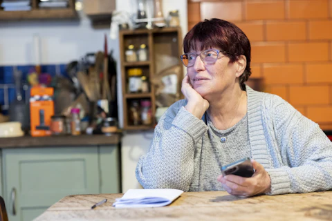 An older woman at her kitchen table, phone in hand, thinking something over